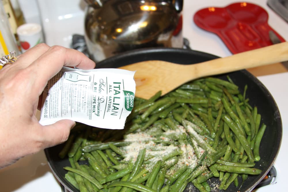 Sautéed green beans in a black fry pan with Good Seasons Italian dressing mix being added