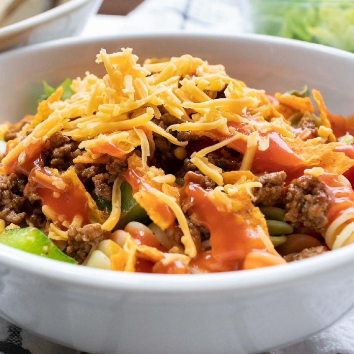 Bowl of seasoned ground beef, spiral pasta, lettuce, halved tomatoes, nacho cheese Doritos, and French salad dressing with shredded cheddar cheese on top 