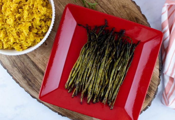 grilled asparagus on a red serving plate placed on a wood serving tray with a bowl of yellow rice and a tea towel to the side