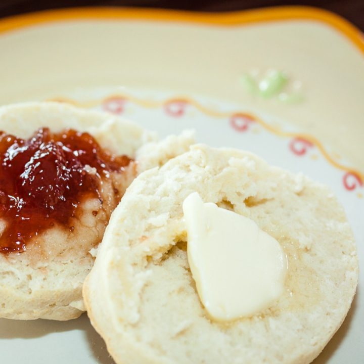 Biscuit cut in half with butter on one half and fruit preserves on the other half on a yellow and white plate