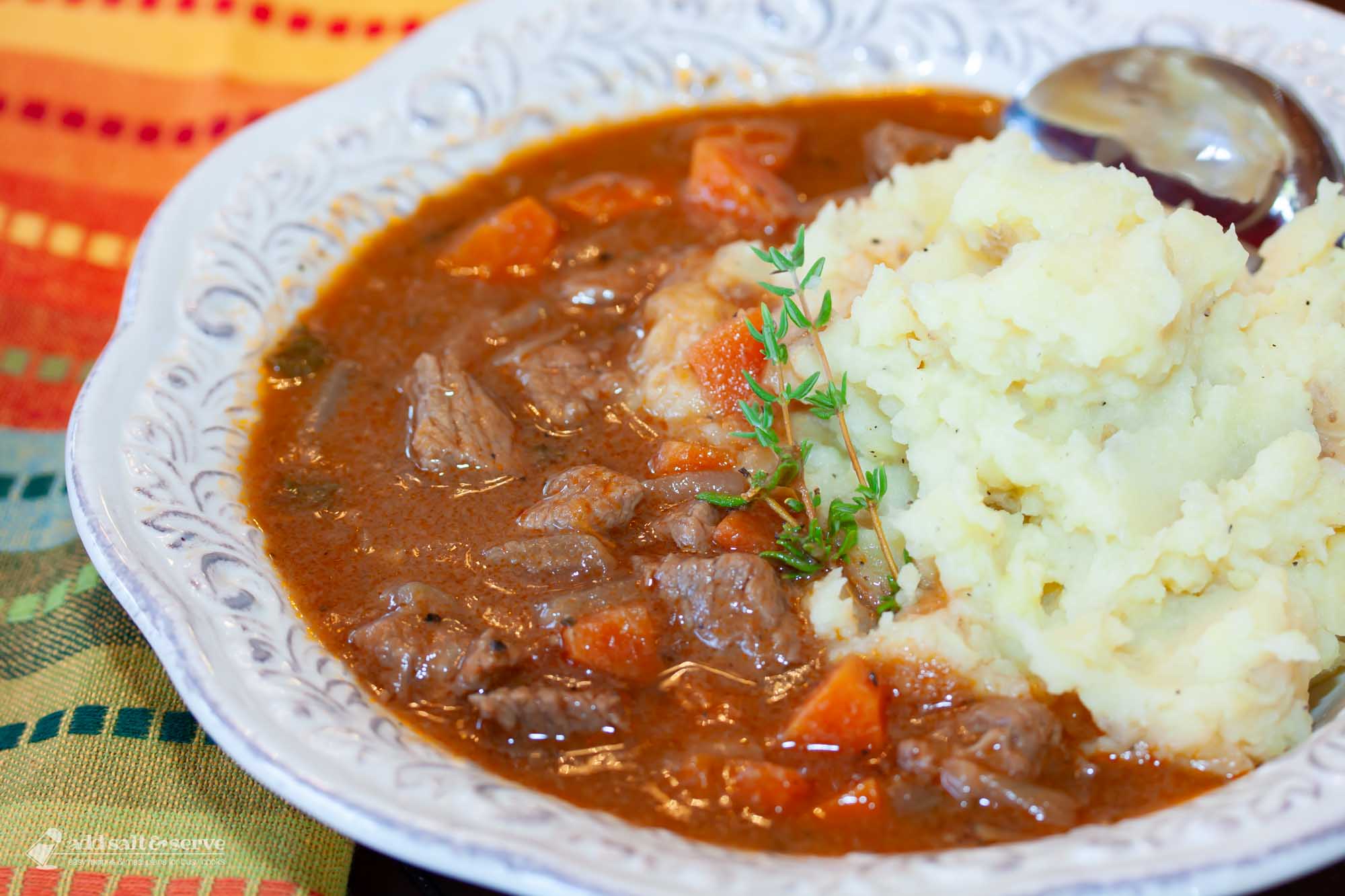 White bowl with beef stew with carrots served over mashed potatoes garnished with a sprig of thyme.