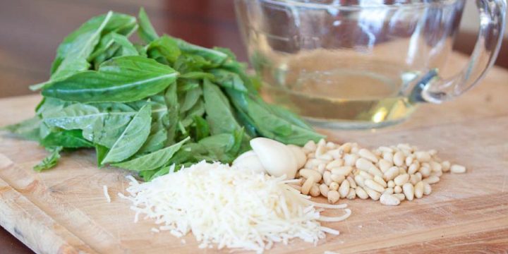 Photo is a pile of basil leaves, a glass measuring cup with olive oil, a pile of shredded parmesan cheese, garlic cloves, and a pile of pine nuts, on a wooden cutting board.