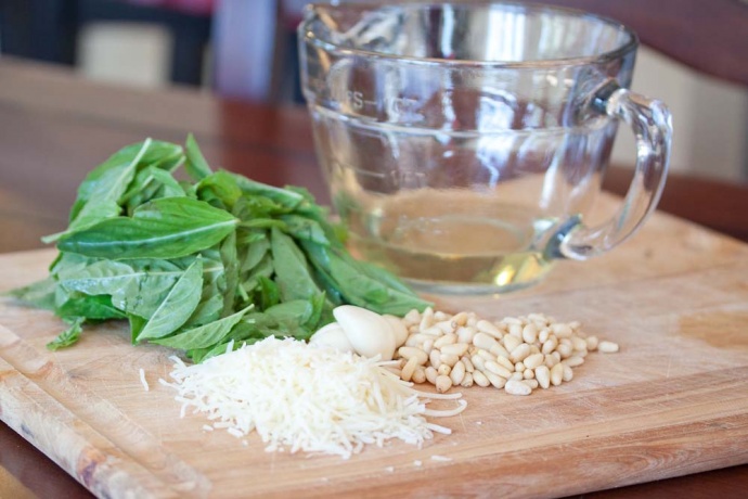 a cutting board with a pile of basil leaves, cloves of garlic, a pile of pine nuts, a pile of shredded Parmesan cheese, and a measuring cup with oil