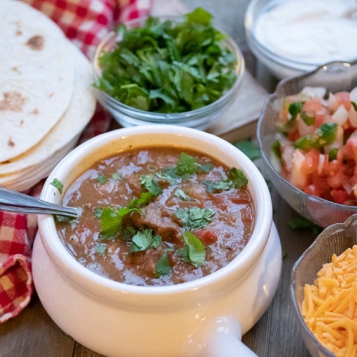 Carne Guisada (Mexican Beef Stew) in a white crock with cheese, tortillas, fresh cilantro, sour cream, and pico de gallo