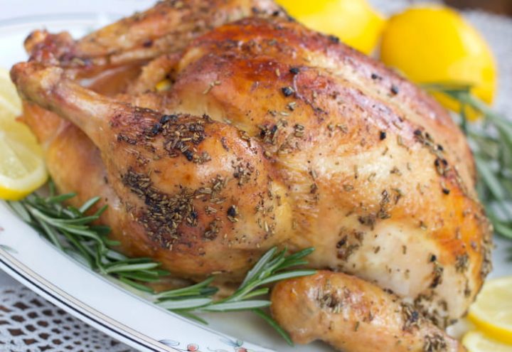 Roasted whole chicken on a white plate, garnished with rosemary and sliced lemon.