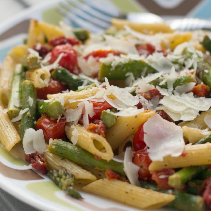 Cooked penne pasta, 1-inch pieces of asparagus, grape tomatoes, topped with a garnish of parmesan cheese on a white plate with blue and brown stripes