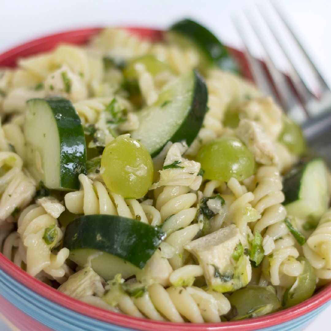 Bowl full of spiral pasta, diced chicken, grapes cut in half, and cucumber slices. There is a fork in the bowl.