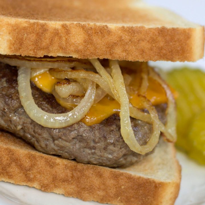 A hamburger patty topped with a slice of melted cheese and sauteed onion rings between two slices of toasted sourdough bread. The sandwich is on a white plate and garnished with pickle slices.