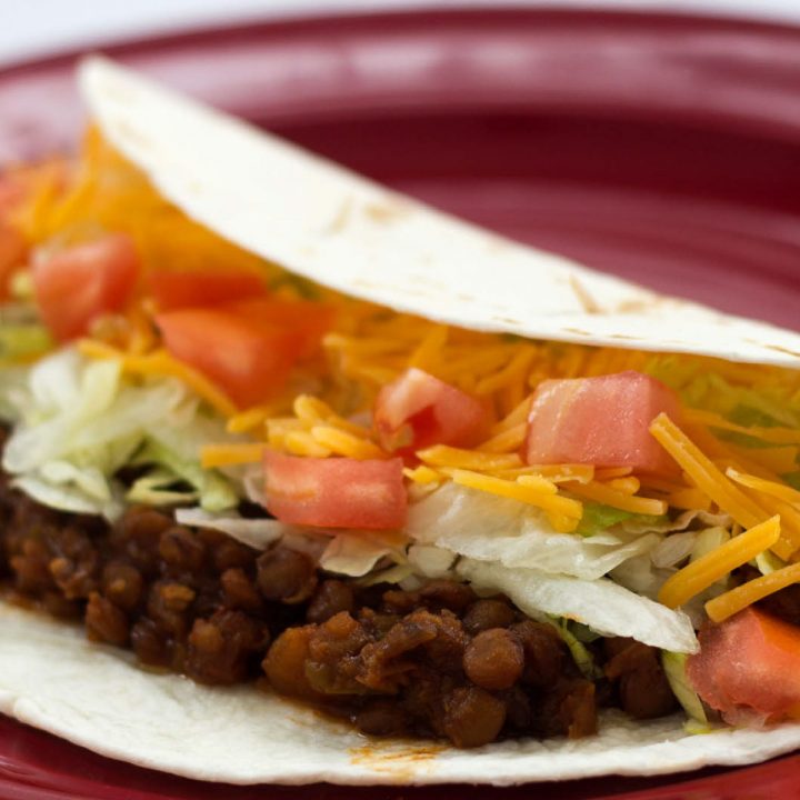 Folded tortilla with lentils, shredded lettuce, diced tomatoes, and shredded cheddar cheese on a red plate.