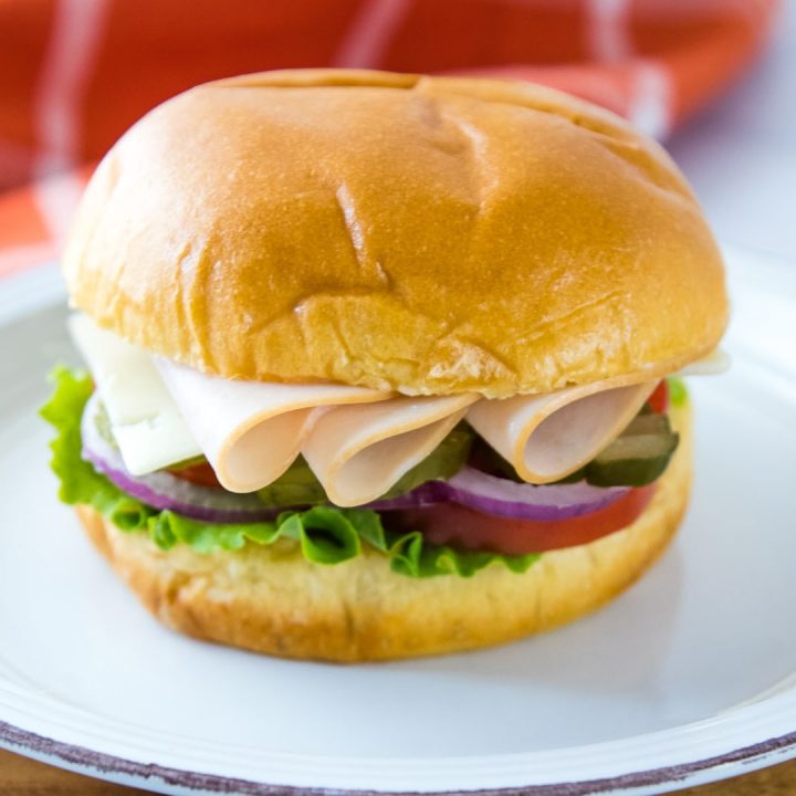 Brioche bun with slices of turkey, Swiss cheese, lettuce, tomato, pickle, and onion on a white and orange plate with coral and white windowpane towel in the background
