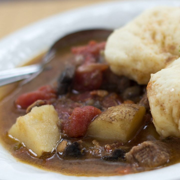 White bowl of beef stew with two dumplings on top. Stew consists of brown broth, diced beef, chopped potatoes, and stewed tomatoes. There is a spoon in the bowl on the left side.