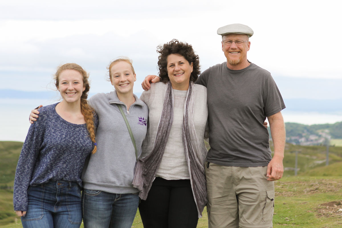 Family with mother, father, and two grown daughters on a hillside with water in background