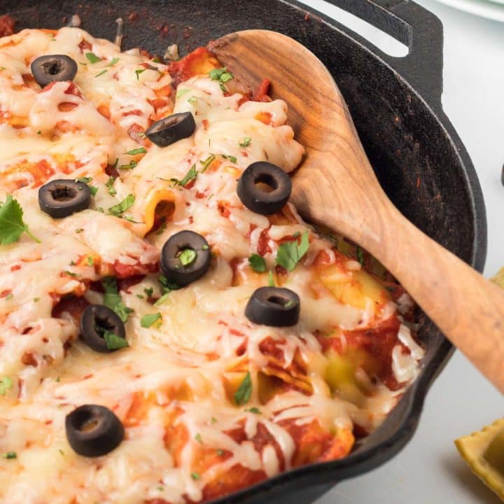 Cast iron skillet with cooked ravioli mixed with salsa, herbs, and tomato sauce and topped with cheese, black olives, and cilantro.