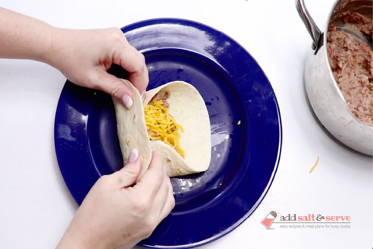 A tortilla with bean burrito filling and shredded cheddar cheese being folded into a burrito.
