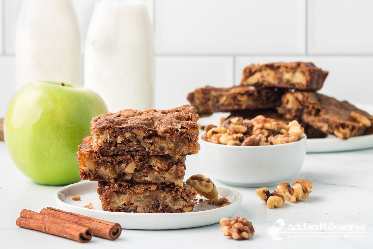 Three Apple Walnut Bars stacked on a white plate with more bars on a plate, a bowl of walnuts, and a green apple in the background.