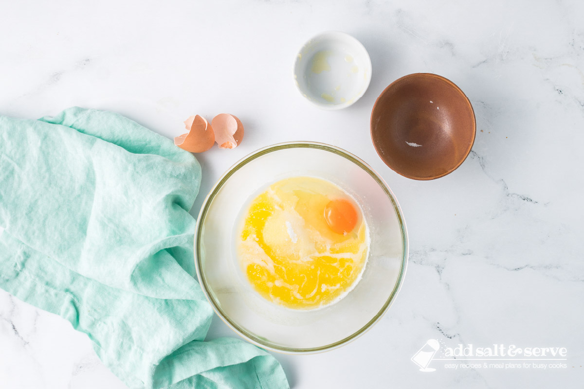 A clear glass bowl with melted butter, a cracked egg, and sugar inside.