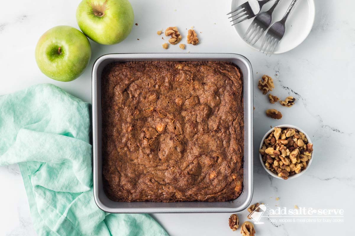 Baked Apple Walnut Bars in an 9-inch by 9-inch pan on a counter surrounded by two green apples, a bowl of walnuts, and a white plate with three forks.