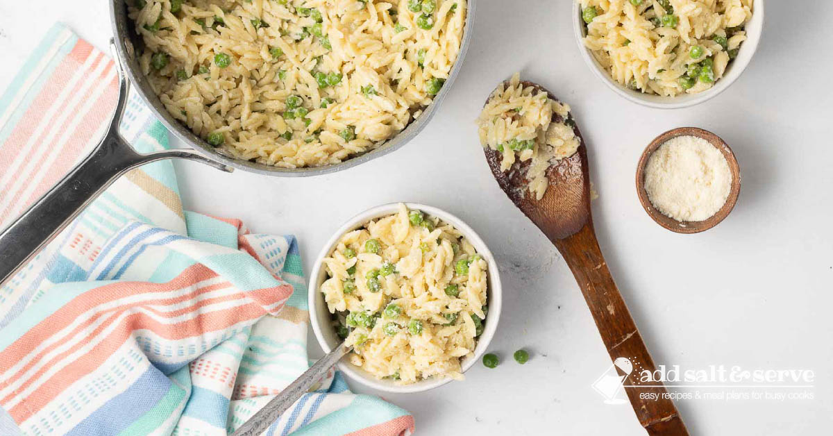 A bowl of Orzo Alfredo with peas with a fork beside the pot of the dish with a wooden spoon laying beside it.