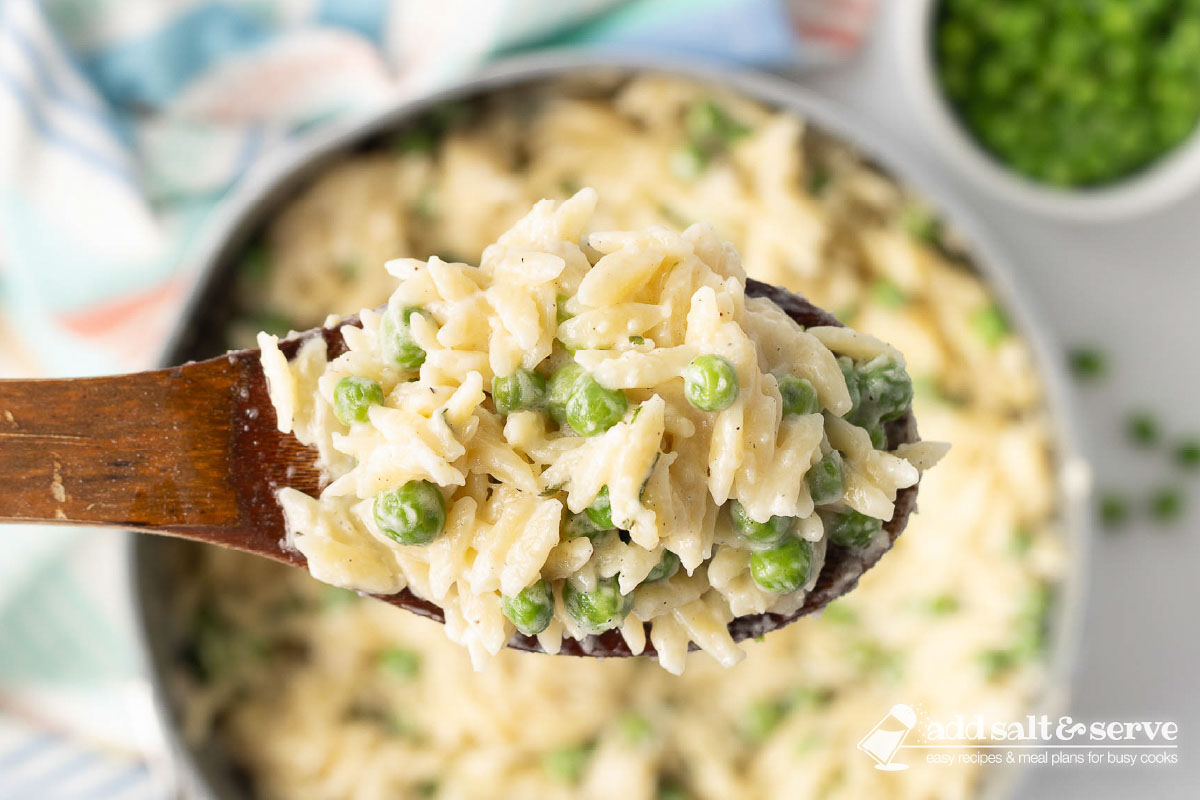 Serving of Orzo Alfredo with peas on a wooden spoon above the pot.