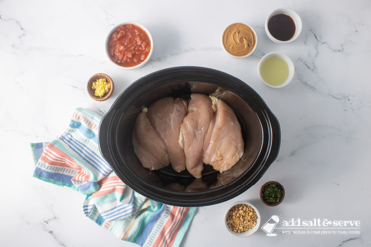 Uncooked chicken breasts in a slow cooker surrounded by small bowls of chopped peanuts, cilantro, peanut butter, salsa, soy sauce, grated ginger, and lime juice.