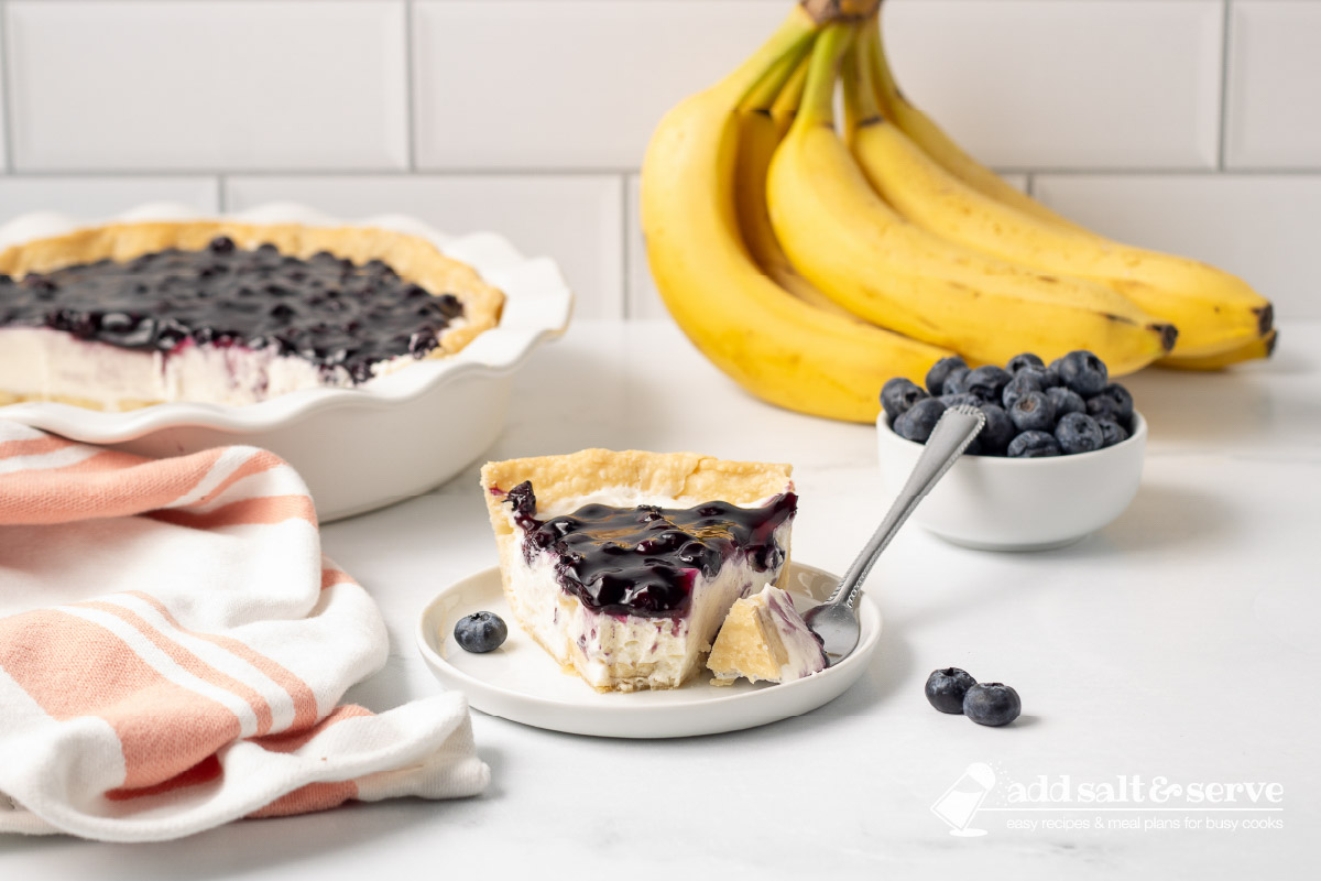 Slice of Blueberry Banana Pie with Cream Cheese with on a plate with a bite on a fork and the sliced pie in the background.