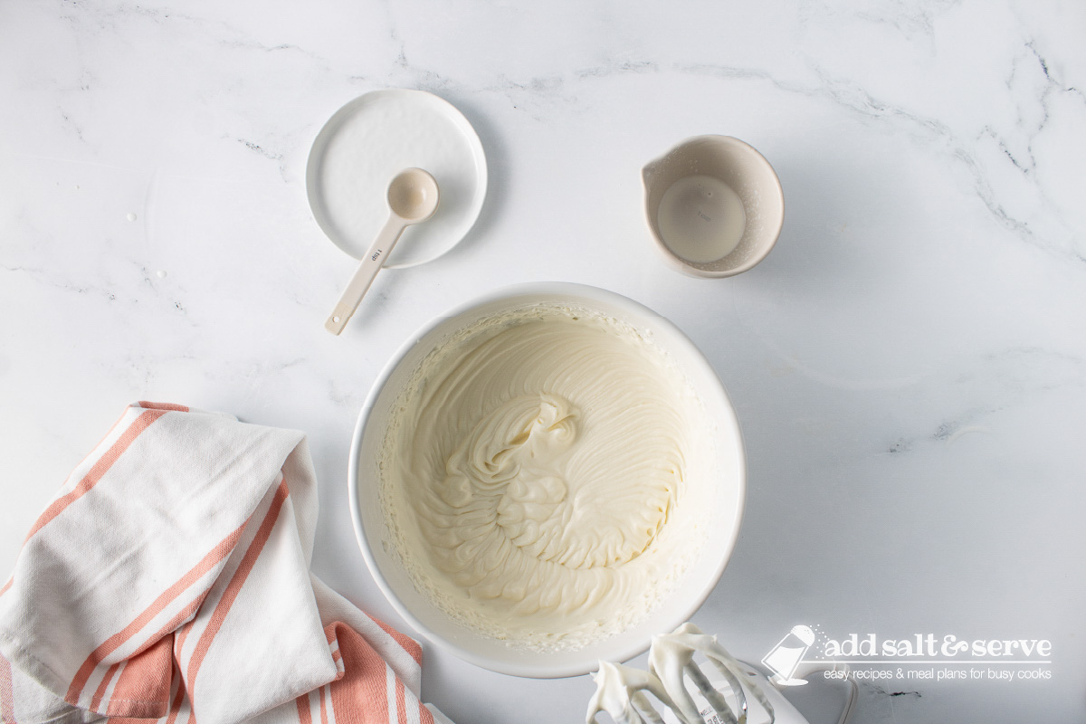 White mixing bowl with whipped cream cheese, sugar, cream, and vanilla extract mixture with empty dishes to the side.