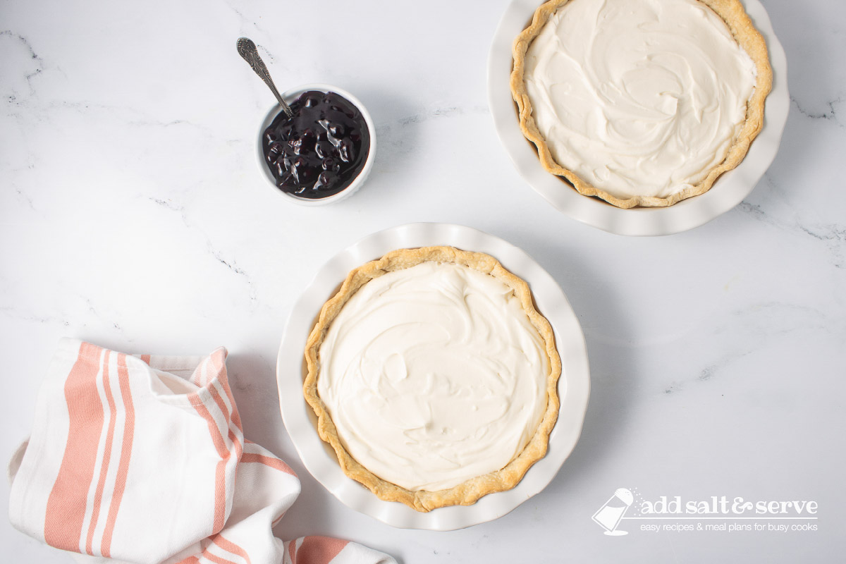 Two pie crusts full of cream cheese mixture with a bowl of blueberry pie filling to the side.