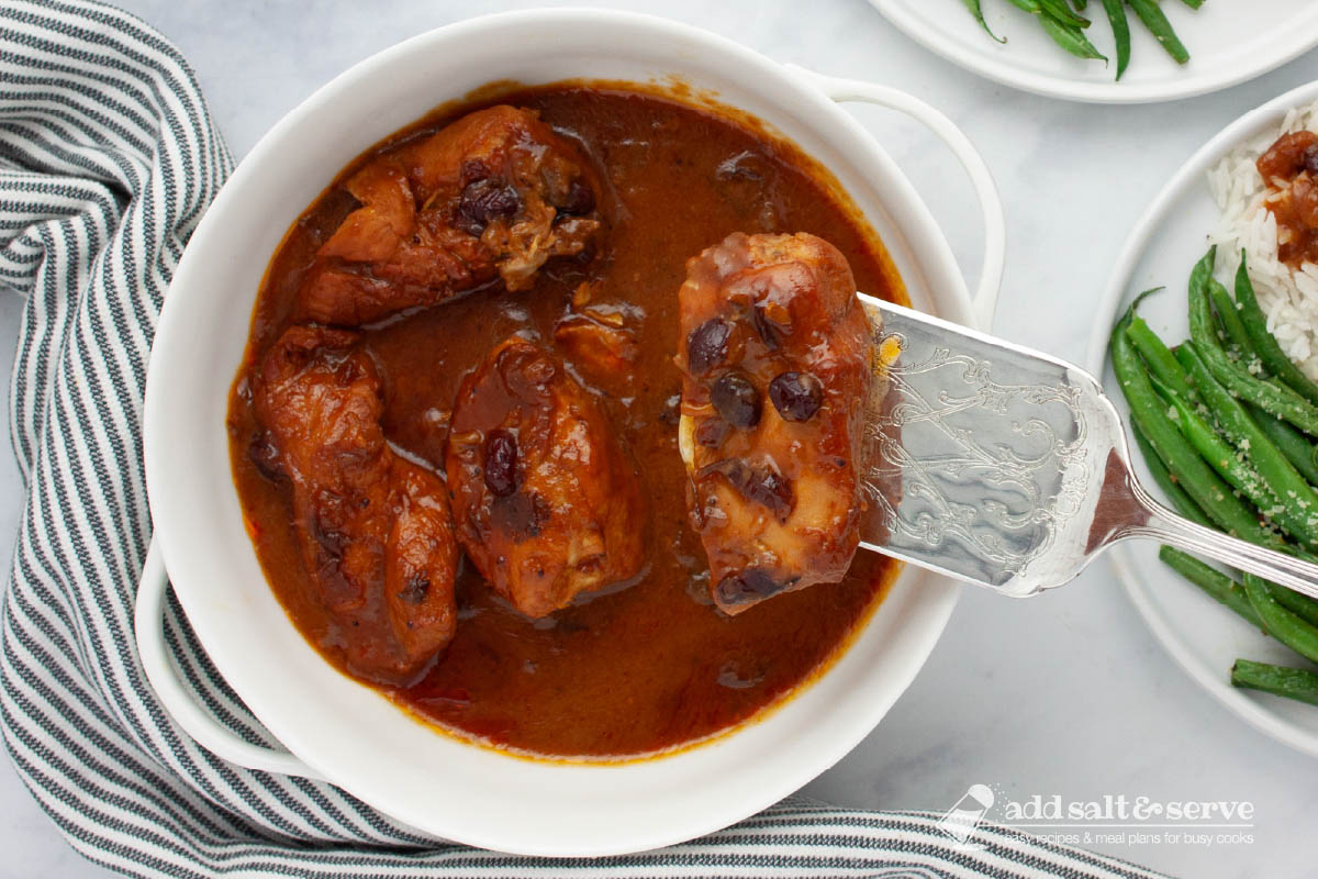 White serving dish with cooked chicken breasts in a sauce made from cranberry sauce with whole cranberries, French dressing, and onion soup mix with a single piece of chicken lifted on a spatula.