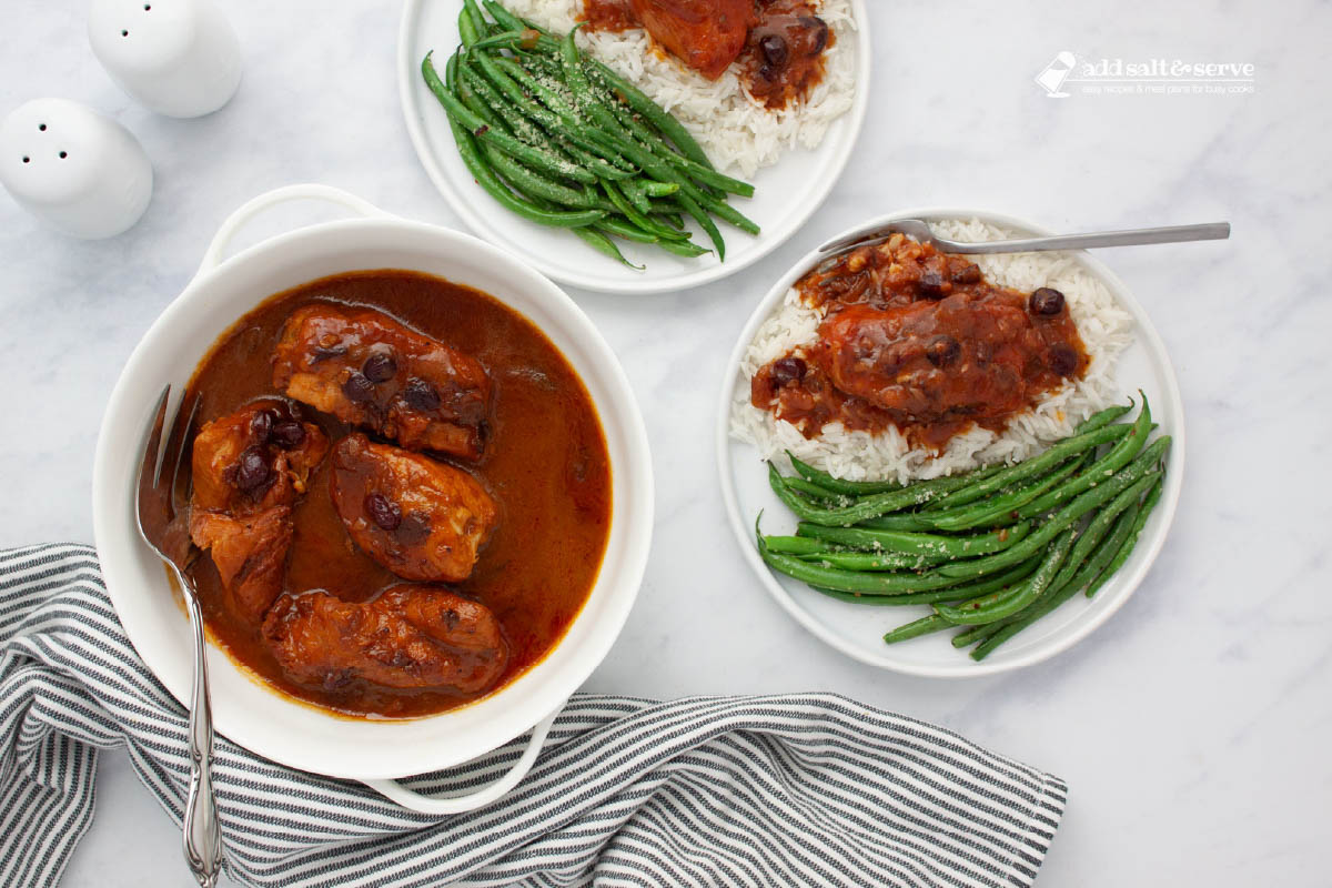 White serving dish with cooked chicken breasts in a sauce made from whole berry cranberry sauce, French dressing, and onion soup mix with a serving fork beside two plates with seasoned green beans and the chicken and sauce over rice on a counter.