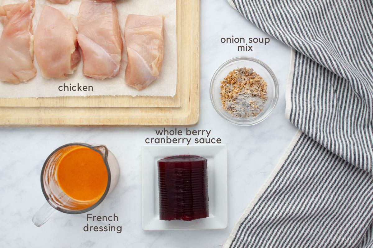 Pieces of uncooked boneless, skinless chicken breast on a cutting board, a measuring cup with French dressing, whole berry cranberry sauce on a plate, and a small bowl of onion soup mix on a counter with a grey striped tea towel to the side.