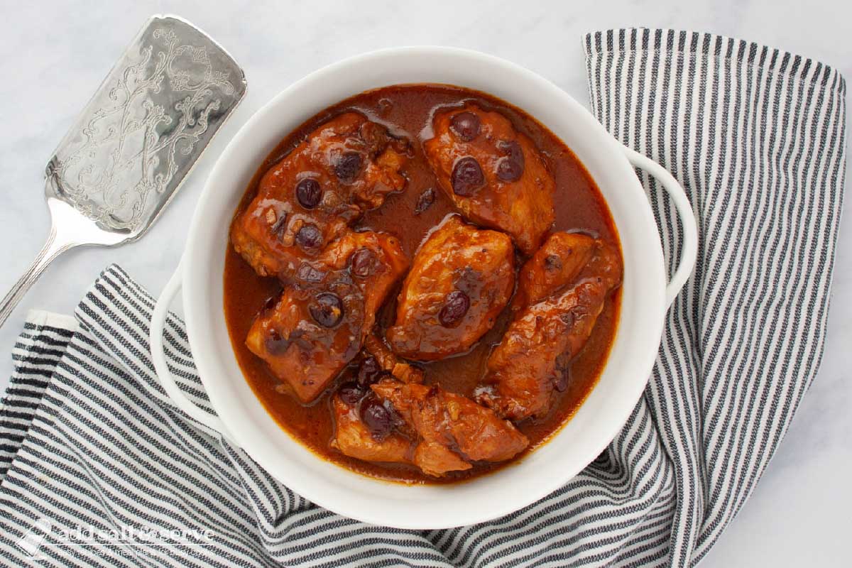 White serving dish with cooked chicken breasts in a sauce made from cranberry sauce with whole cranberries, French dressing, and onion soup mix beside a grey striped tea towel and a silver spatula.