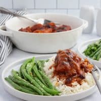 A white plate with seasoned green beans and chicken in a reddish-orange sauce with cranberries served over white rice with a serving dish of the chicken in the background.