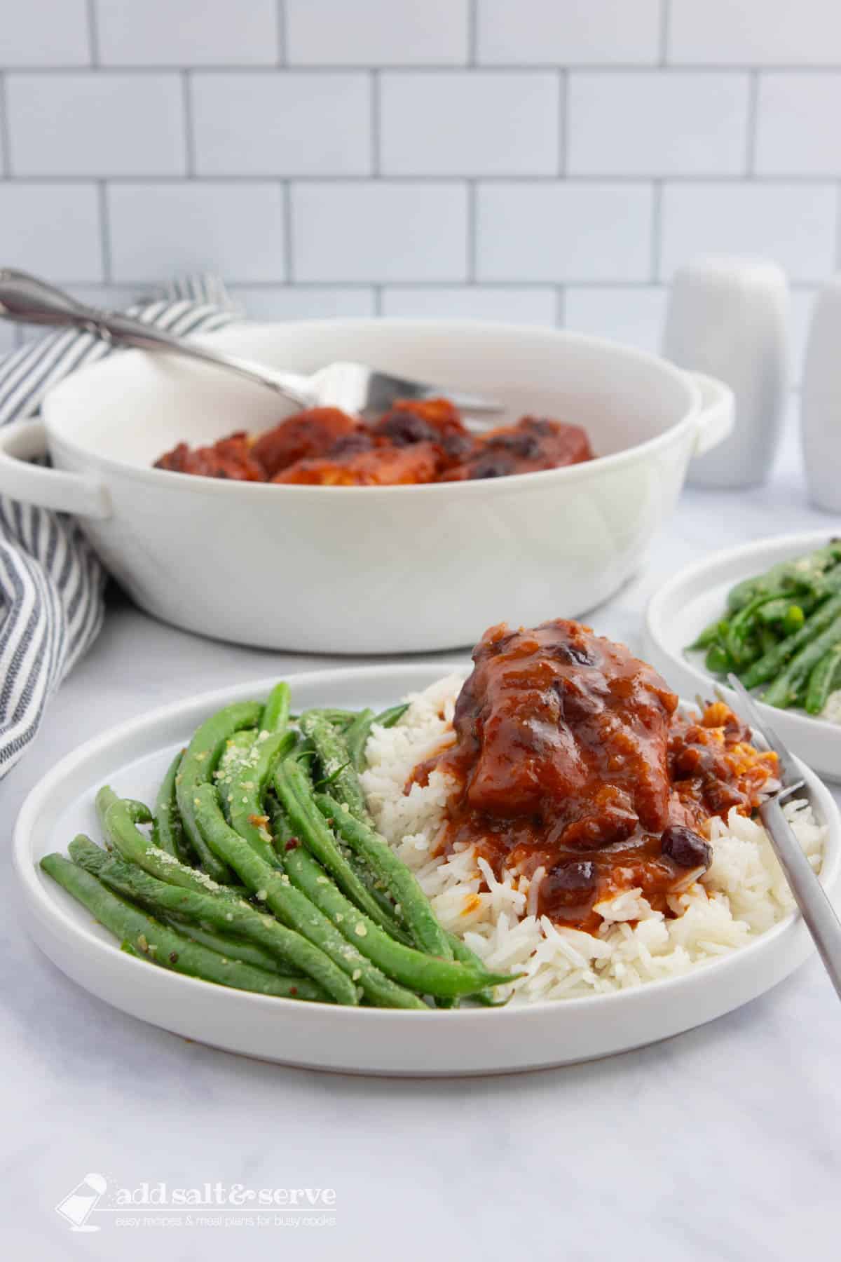 A white plate with seasoned green beans and chicken in a reddish-orange sauce with cranberries served over white rice with a serving dish of the chicken in the background.