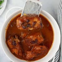 White serving dish with cooked chicken breasts in a sauce made from cranberry sauce with whole cranberries, French dressing, and onion soup mix with a single piece of chicken lifted on a spatula.