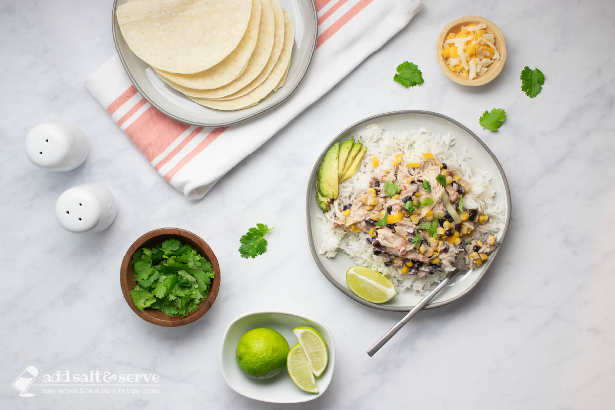 A plate of shredded chicken in a creamy sauce with black beans, corn, tomatoes, and green chilies served over rice with garnishes of sliced avocado, shredded cheese, fresh cilantro, and lime wedges and a plate of corn tortillas.