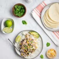 A plate of shredded chicken in a creamy sauce with black beans, corn, tomatoes, and green chilies served over rice and garnished with sliced avocado, shredded cheese, fresh cilantro, and lime wedges beside a plate of corn tortillas, a bowl of shredded cheese, a bowl of fresh cilantro, and a bowl with lime wedges.