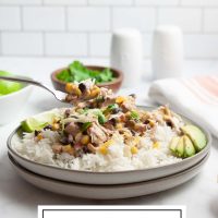 A plate of shredded chicken in a creamy sauce with black beans, corn, tomatoes, and green chilies served over rice and garnished with sliced avocado, shredded cheese, fresh cilantro, and lime wedges beside a bowl of fresh cilantro and a bowl with lime with text Crock Pot Fiesta Chicken.