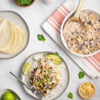 A plate of shredded chicken in a creamy sauce with black beans, corn, tomatoes, and green chilies served over rice and garnished with sliced avocado, shredded cheese, fresh cilantro, and lime wedges beside a bowl of the shredded chicken in sauce, a plate of corn tortillas, a bowl of shredded cheese, a bowl of fresh cilantro, and a bowl with lime wedges.