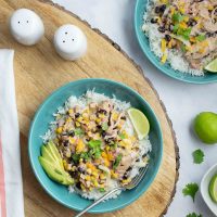 Bowl of shredded chicken in a creamy sauce with black beans, corn, tomatoes, and green chilies over rice and garnished with sliced avocado, shredded cheese, fresh cilantro, and lime wedges.