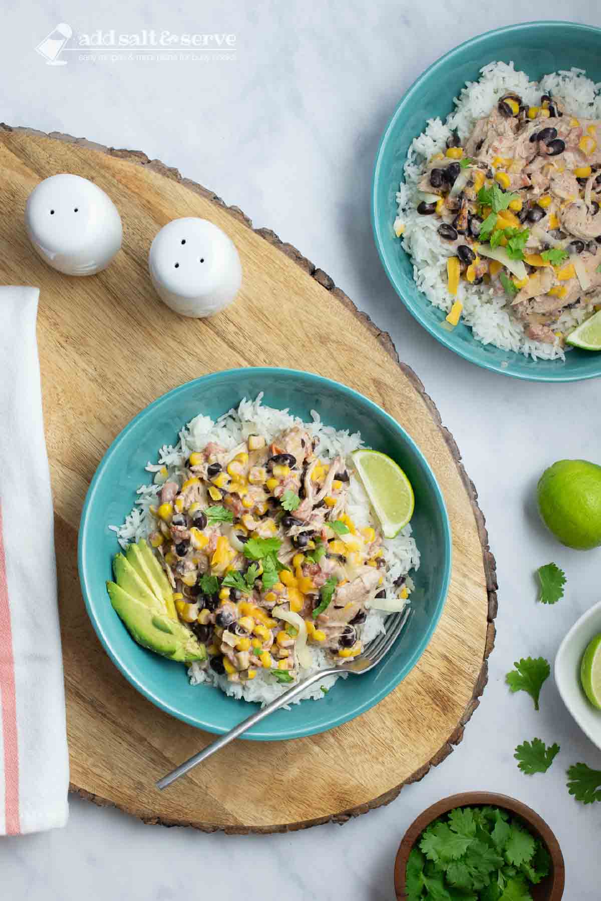 Bowl of shredded chicken in a creamy sauce with black beans, corn, tomatoes, and green chilies over rice and garnished with sliced avocado, shredded cheese, fresh cilantro, and lime wedges.