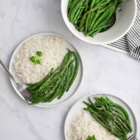 Seasoned green beans garnished with Parmesan cheese on plates with white rice beside a serving bowl of seasoned green beans.