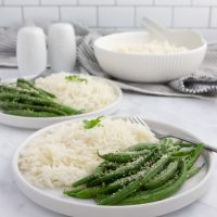 Seasoned green beans garnished with Parmesan cheese on plates with white rice with a serving bowl of rice in the background.