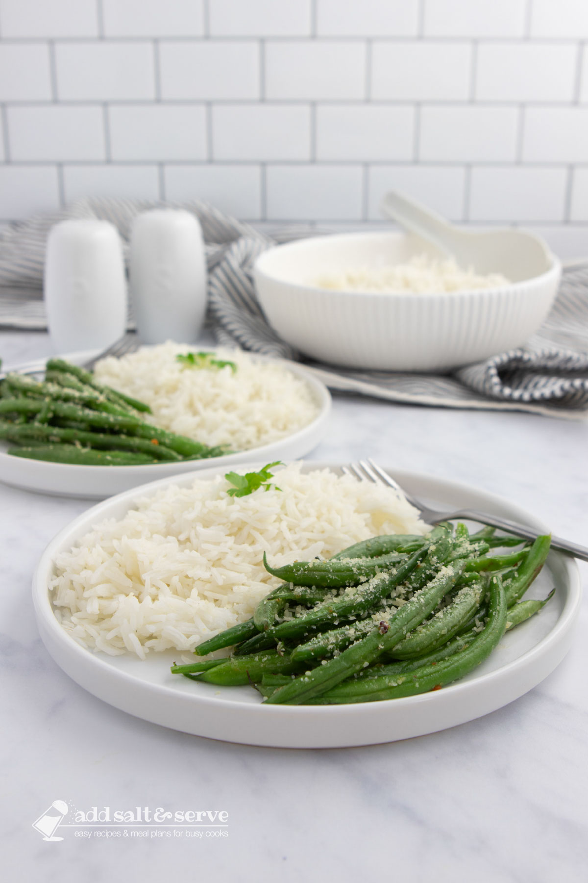 Seasoned green beans garnished with Parmesan cheese on plates with white rice with a serving bowl of rice in the background.