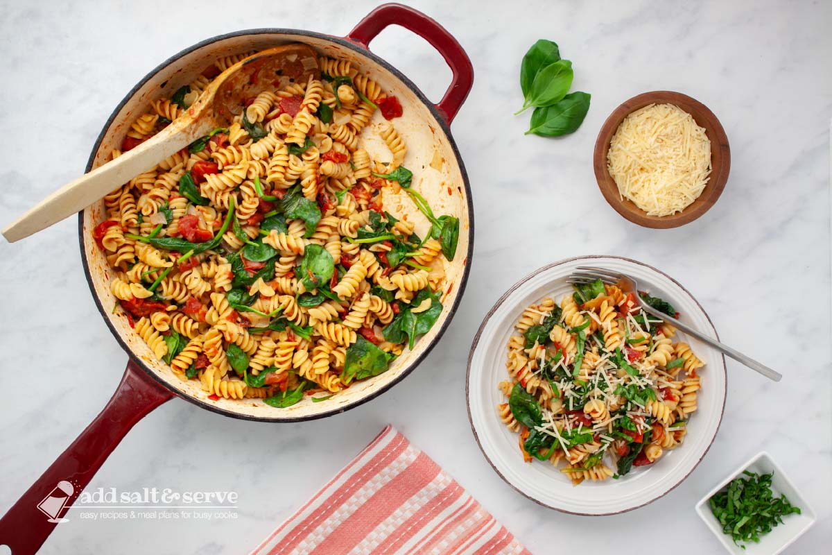 Plate of tomato Spinach Pasta garnished with Parmesan cheese and fresh basil strips on a counter beside the pan of the full dish.