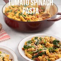 Two plates of Tomato Spinach Pasta garnished with Parmesan cheese and fresh basil strips on a counter beside the pan of the full dish and a small bowl of fresh basil strips with text One Pot Tomato Spinach Pasta.