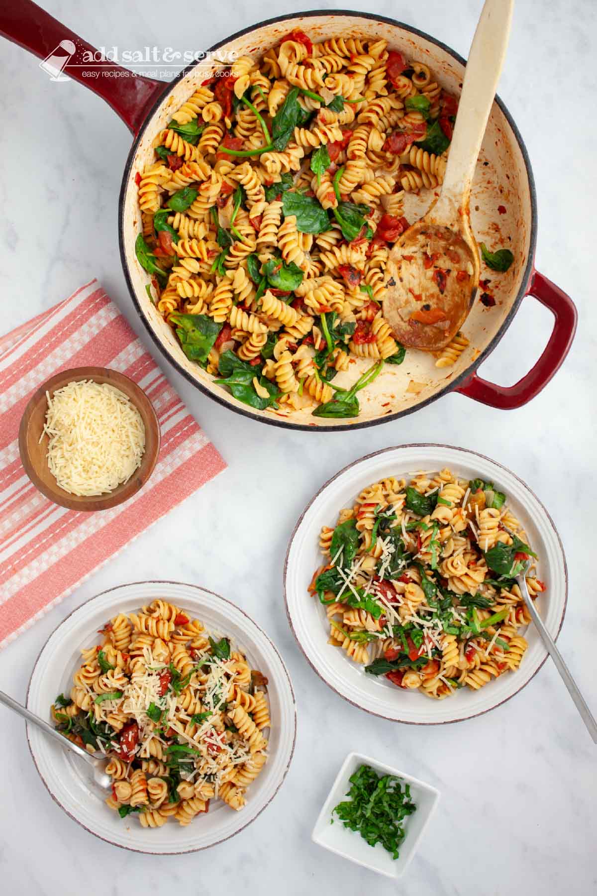 Two plates of tomato Spinach Pasta garnished with Parmesan cheese and fresh basil strips on a counter beside the pan of the full dish and small bowls of parmesan cheese and fresh basil strips.