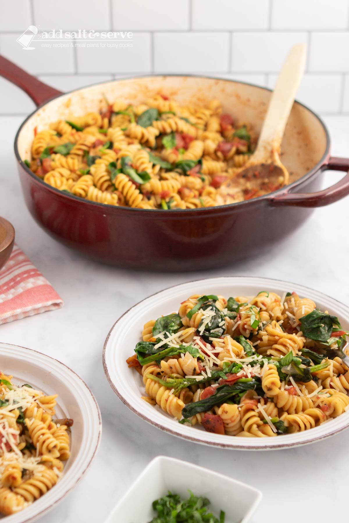 Plate of tomato Spinach Pasta garnished with Parmesan cheese and fresh basil strips on a counter beside the pan of the full dish.