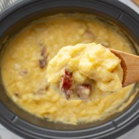 Close-up of a spoonful of cheesy potato soup with kielbasa pieces on a wooden spoon held over a crockpot of the soup.