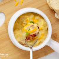 Close-up of a spoonful of cheesy potato soup with kielbasa pieces garnished with shredded cheddar and sliced green onion on a spoon held over a crock of the soup.