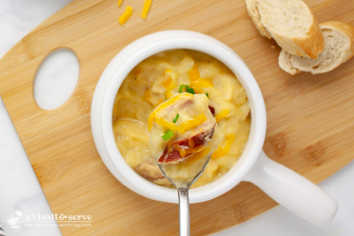 Close-up of a spoonful of cheesy potato soup with kielbasa pieces garnished with shredded cheddar and sliced green onion on a spoon held over a crock of the soup.